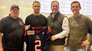 Zwei Männer halten ein Football-Trikot mit der Nummer 2 und stehen neben einem Mann mit einer Halskette und einem Mann mit einer grünen Weste. Ein großer Bildschirm im Hintergrund zeigt Fluginformationen.