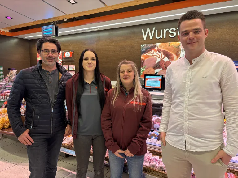 Vier Personen, zwei Männer und zwei Frauen, stehen in einem Supermarkt. Eine Frau hat blonde Haare, die andere dunkle Haare. Hinter ihnen ist eine Fleischtheke mit Produkten.