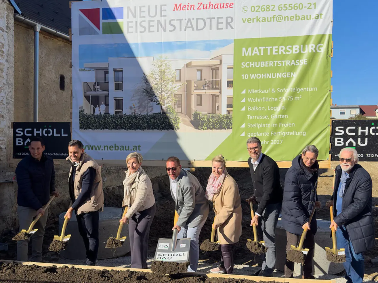 Mehrere Personen stehen an einer Baustelle und halten Schaufeln. Ein grüner Banner zeigt hinter ihnen ein Haus mit der Aufschrift 'Mattersburg Schubertstrasse 10 Wohnungen'. Sie tragen alle Jacken und lächeln.