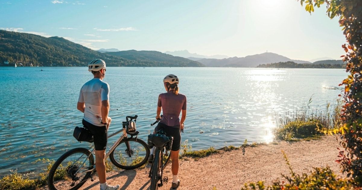Zwei Radfahrer, ein Mann und eine Frau, stehen am Ufer eines Sees mit Bergen im Hintergrund. Die Sonne geht unter und taucht die Szene in ein warmes Licht.