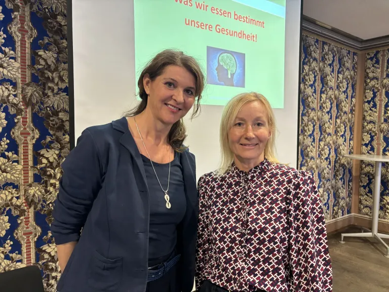 Zwei Frauen stehen nebeneinander vor einer Präsentationsleinwand, die ein Bild eines Gehirns mit der Überschrift 'Was wir essen, bestimmt unsere Gesundheit!' zeigt.