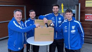 Vier Männer in blauen Jacken posieren mit einer Box mit der Aufschrift Sammelbox. Die Box ist braun mit grünem Text, und der Mann auf der rechten Seite hält ein Stück Papier.