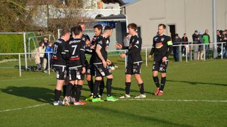Fußballspieler in schwarzen Trikots versammeln sich auf einem Feld und unterhalten sich. Ein Ball liegt in der Nähe auf dem Boden.