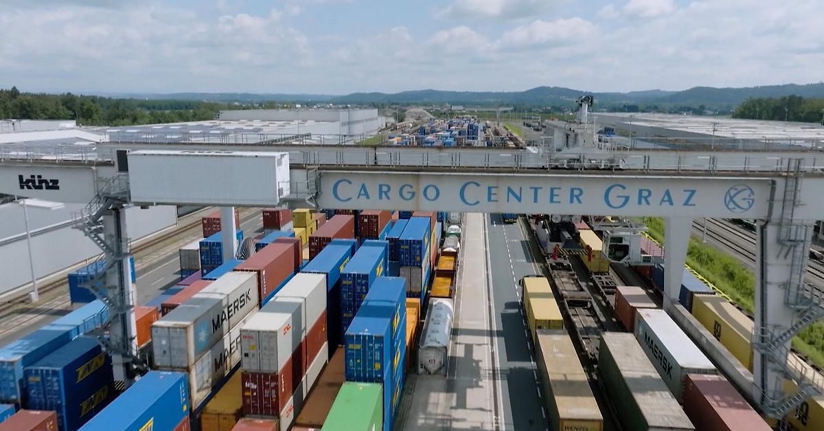 Luftaufnahme eines Frachtzentrums mit gestapelten Containern. Das Schild oben liest sich 'CARGO CENTER GRA'. Viele LKWs und Züge sind geparkt, Kräne sind sichtbar.