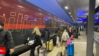Eine Gruppe Reisender wartet an einem Bahnhof in der Nähe eines roten Zuges mit dem Wort flix an der Seite. Sie tragen verschiedene farbiges Gepäck und tragen Winterkleidung.