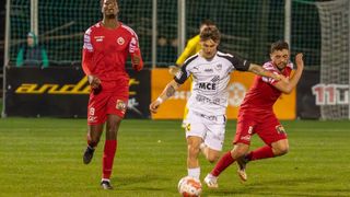 Fußballspieler in roten und weißen Trikots spielen auf einem Feld. Ein Spieler läuft auf den Ball zu, ein anderer verteidigt. Ein Werbebanner ist im Hintergrund zu sehen.