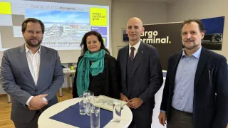 Vier Personen stehen lächelnd vor einem Tisch mit Gläsern, wahrscheinlich bei einer Veranstaltung. Eine Projektionsleinwand hinter ihnen zeigt eine Präsentation.