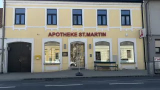Die Vorderseite eines gelben Gebäudes mit dem Schild 'APOTHEKE ST.MARTIN' über dem Eingang. Davor steht eine Bank und ein Auto ist geparkt.