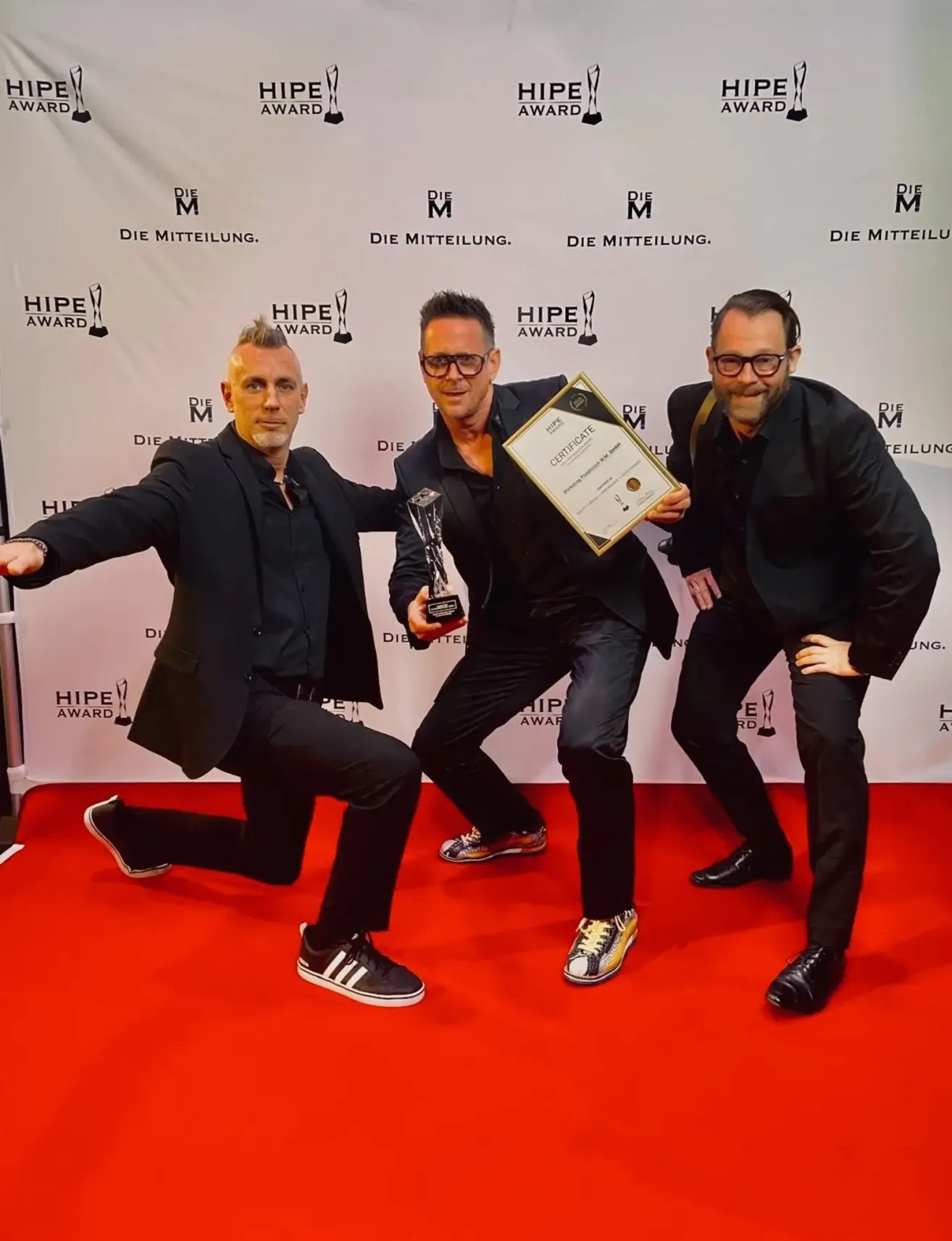 Drei Männer in schwarzen Anzügen posieren auf einem roten Teppich, einer hält eine Trophäe und ein Zertifikat. Hinter ihnen ist ein weißer Hintergrund mit dem Text 'DIE MITTEILUNG' und 'HIPE AWARD'.