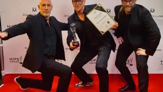 Drei Männer in schwarzen Anzügen posieren auf einem roten Teppich, einer hält eine Trophäe und ein Zertifikat. Hinter ihnen ist ein weißer Hintergrund mit dem Text 'DIE MITTEILUNG' und 'HIPE AWARD'.