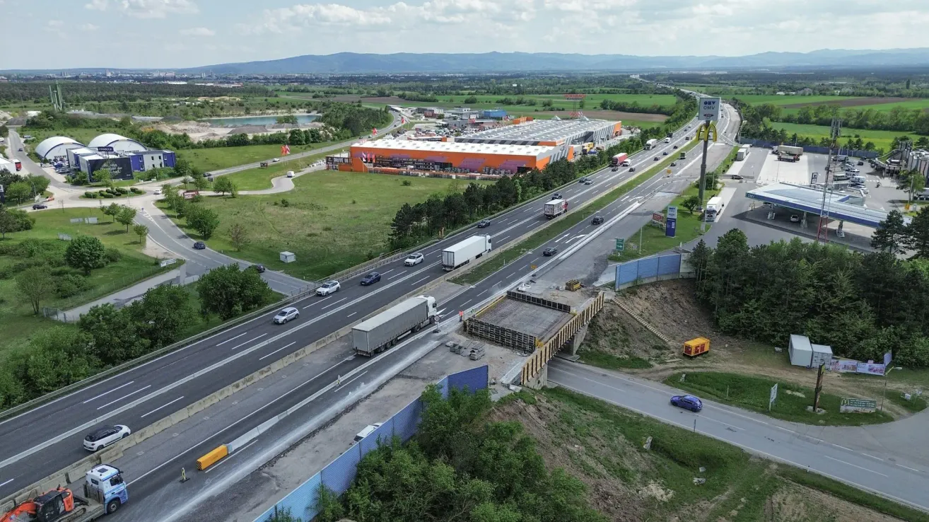 Ein Luftbild einer Autobahn mit einer Brücke im Bau und einem großen Gebäude in der Ferne. Viele Autos und LKWs sind auf der Straße.
