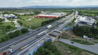 Ein Luftbild einer Autobahn mit einer Brücke im Bau und einem großen Gebäude in der Ferne. Viele Autos und LKWs sind auf der Straße.
