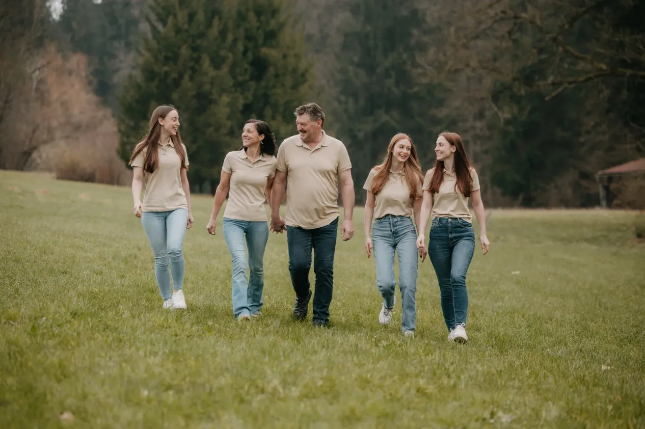 Eine fünfköpfige Familie läuft lächelnd und Hand in Hand auf einer Grasfläche. Sie tragen passende beigefarbene Hemden und blaue Jeans. Im Hintergrund sind Bäume zu sehen.
