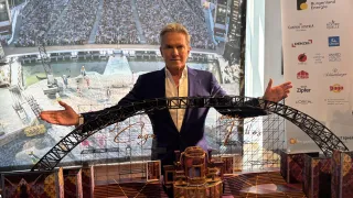 Ein Mann steht vor einem Modell einer Showbühne mit weit ausgebreiteten Armen. Das Modell der Bühne ist detailliert mit einem goldenen Käfig in der Mitte und einem Bogen dahinter. Hinter dem Modell ist eine große Leinwand zu sehen, die eine Menge zeigt.