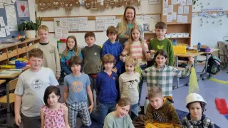Ein Klassenzimmer voller Kinder mit einer Frau in der Mitte. Die Kinder sitzen und knien auf dem blauen Teppich. Es gibt Schreibtische und Stühle.