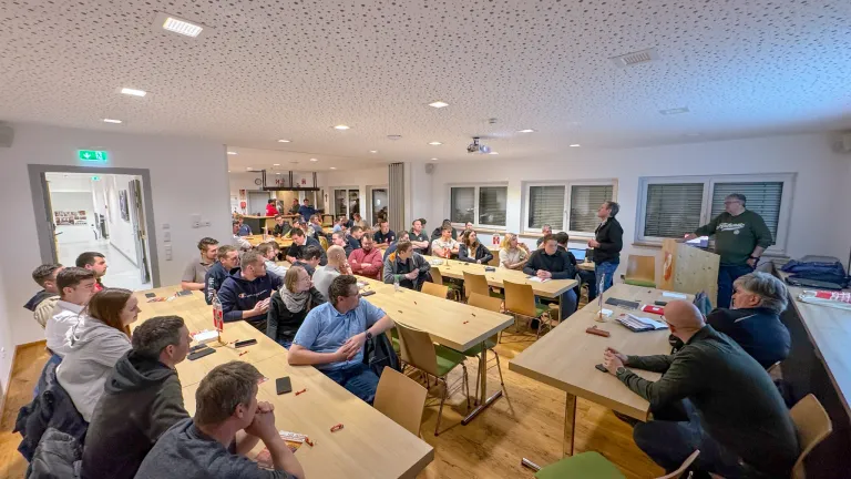 Ein Seminarraum mit Menschen an Tischen. Eine Person steht vorne und spricht die Teilnehmer an. Der Raum hat einen Holzboden, weiße Wände und Fenster mit Jalousien.