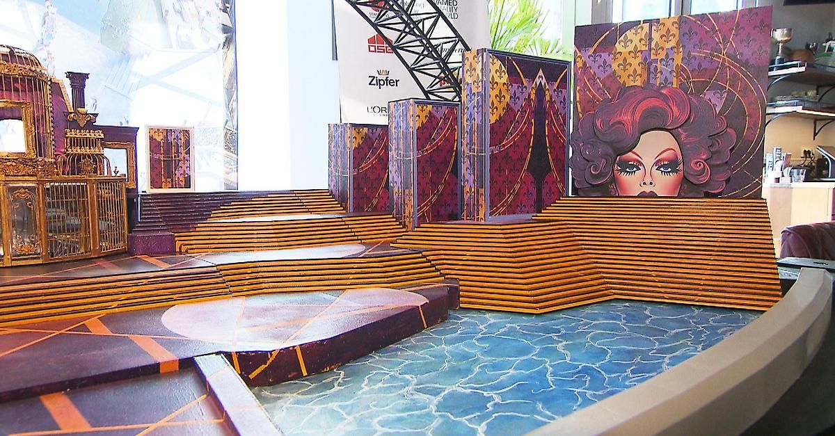 Eine farbenfrohe Darstellung einer Bühnenausstattung mit gelben Stufen, geschmückt mit farbenfrohen Kulissen, darunter eine mit dem Gesicht einer Frau. Der Boden hat eine blaue, wellenförmige Textur, und es gibt Werbung auf Bannern im Hintergrund.