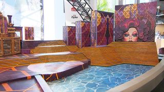 Eine farbenfrohe Darstellung einer Bühnenausstattung mit gelben Stufen, geschmückt mit farbenfrohen Kulissen, darunter eine mit dem Gesicht einer Frau. Der Boden hat eine blaue, wellenförmige Textur, und es gibt Werbung auf Bannern im Hintergrund.