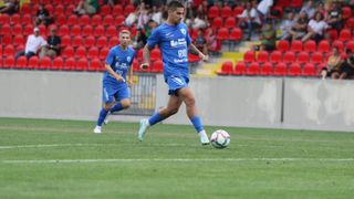 Zwei Fußballspieler in blauen Trikots spielen auf einem Feld. Einer dribbelt den Ball, während der andere hinter ihm läuft. Dahinter schaut ein Publikum aus roten Tribünenplätzen zu.