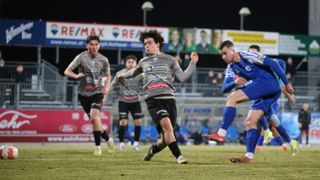 Ein Nachtfußballspiel mit Spielern in grauen, schwarzen und blauen Trikots. Ein Spieler hebt die Faust, und ein anderer versucht zu blocken. Zuschauer sitzen im Hintergrund.