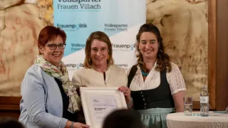 Drei Frauen stehen vor einer Kulisse mit dem Volkspartei Frauen Villach-Banner, lächeln und halten ein gerahmtes Zertifikat. Eine Frau trägt eine blaue Halskette, eine andere einen Blumenschal. Ein Tisch mit einer Wasserflasche und einem Glas steht hinter ihnen.