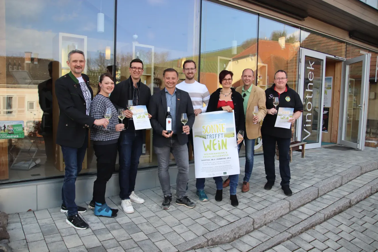 Eine Gruppe von Menschen in Geschäftskleidung posiert vor einem Geschäft mit Weinflaschen und Gläsern. Sie halten ein Plakat, auf dem Sonne trifft Wein steht.