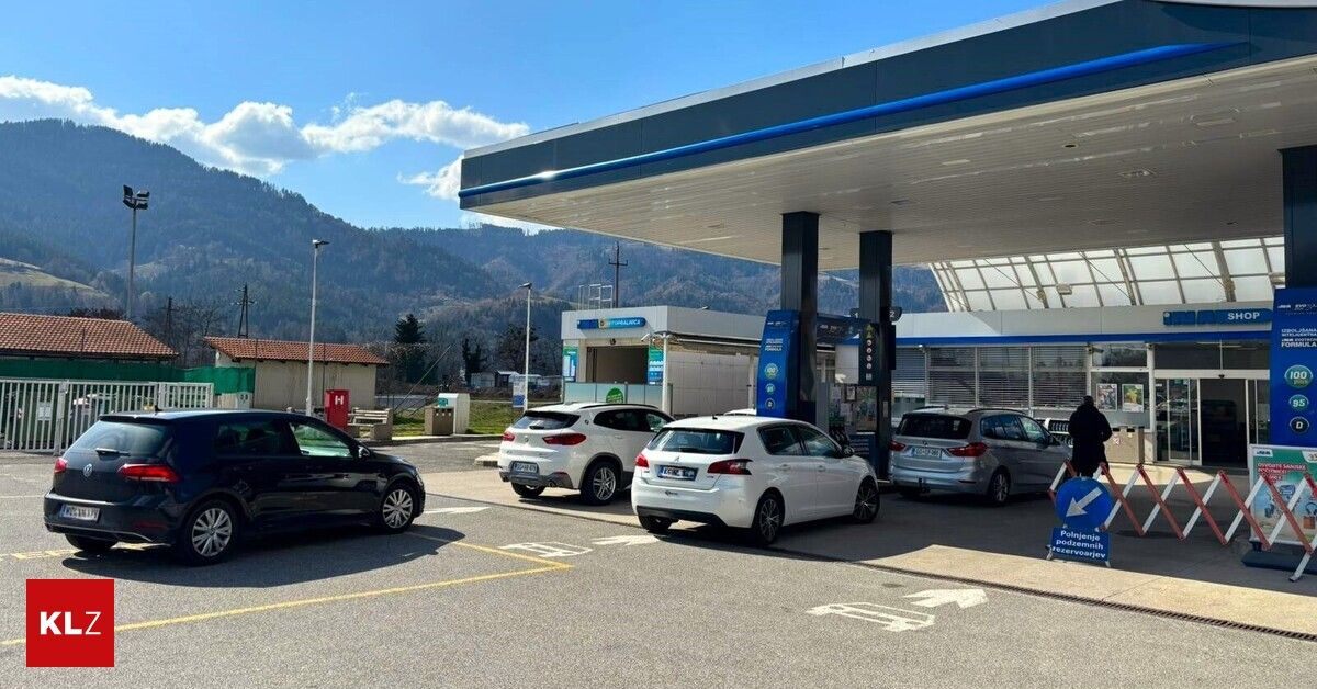 Eine Tankstelle mit mehreren geparkten Autos davor. Die Tankstelle hat ein modernes Design mit blauen und weißen Farben. Im Hintergrund sind Berge sichtbar.