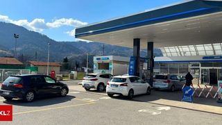 Eine Tankstelle mit mehreren geparkten Autos davor. Die Tankstelle hat ein modernes Design mit blauen und weißen Farben. Im Hintergrund sind Berge sichtbar.