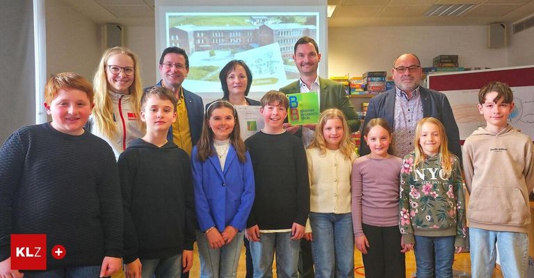 Eine Gruppe von Erwachsenen und Kindern steht zusammen, hält ein grünes Buch und Papier und lächelt in einem Klassenzimmer. Eine Projektionsleinwand zeigt ein Gebäude im Hintergrund.