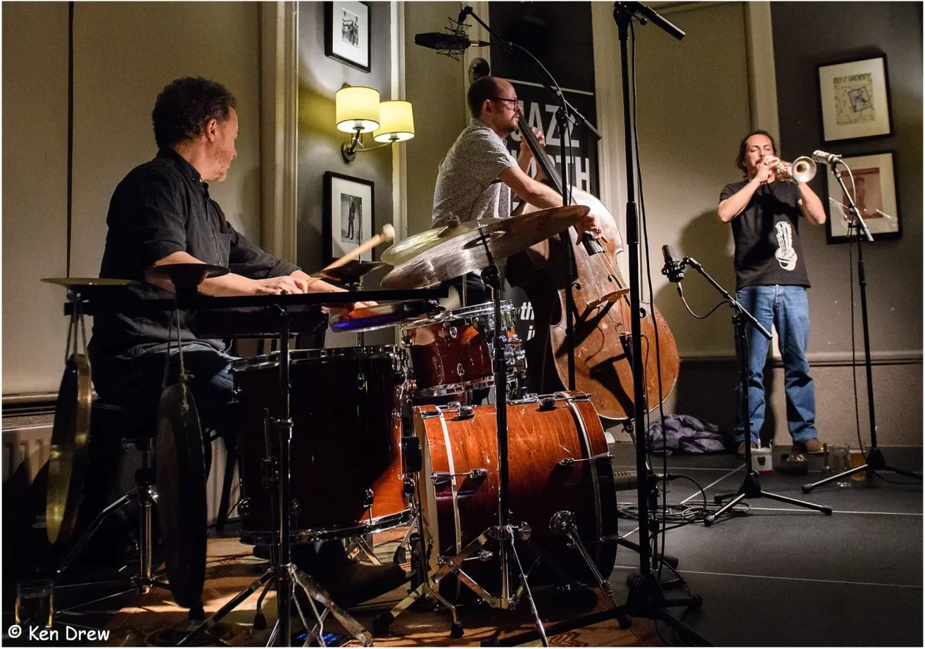 Ein Jazz-Trio tritt auf. Der Schlagzeuger spielt am Schlagzeug, der Bassist hält einen Bogen, und der Trompeter steht vor einem Mikrofon. Der Raum ist schwach beleuchtet.