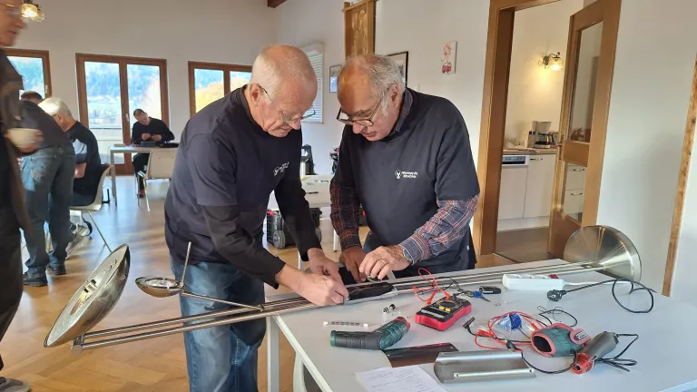 Zwei ältere Männer arbeiten an einem elektronischen Projekt an einem Tisch, umgeben von Werkzeugen und Kabeln. Sie befinden sich in einem Raum mit Holzböden und großen Fenstern.