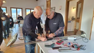 Zwei ältere Männer arbeiten an einem elektronischen Projekt an einem Tisch, umgeben von Werkzeugen und Kabeln. Sie befinden sich in einem Raum mit Holzböden und großen Fenstern.