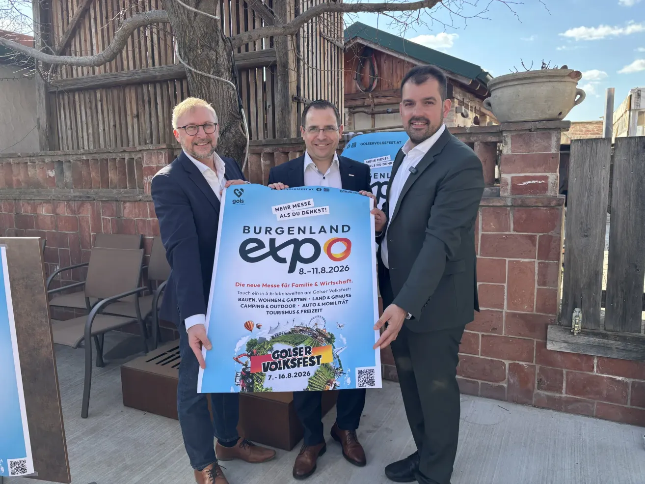 Drei Männer in Anzügen stehen draußen und halten ein großes blaues Schild, das für die Burgenland Expo 2026 wirbt, mit einer lächelnden Frau in der Mitte. Dahinter ist eine Backsteinmauer und ein Baum.