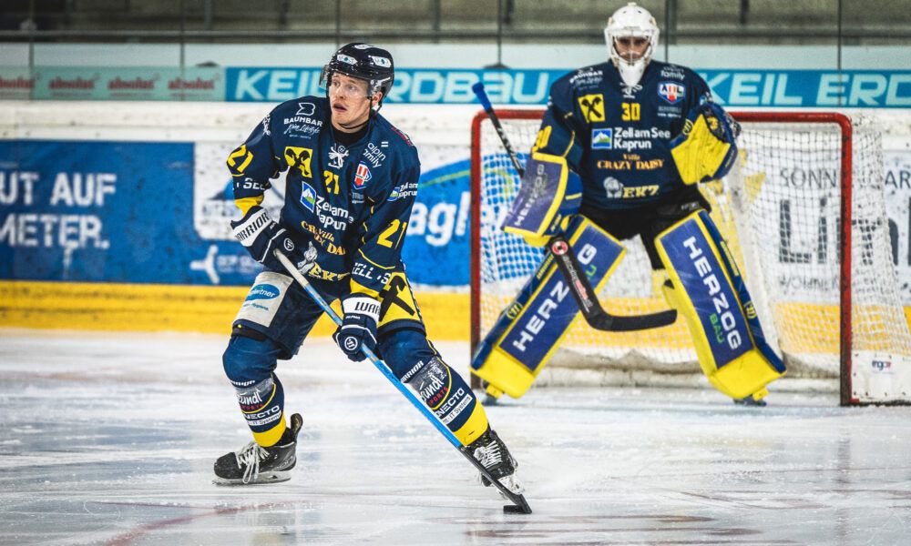 Zwei Eishockeyspieler auf dem Eis, einer mit der Nummer 21 und der andere mit 30, beide tragen Trikots mit Sponsorenlogos. Sie spielen in einer überdachten Arena.