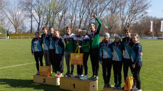 Eine Gruppe junger Athleten steht auf einem Podest mit Medaillen um den Hals. Sie halten eine gelbe Tasche mit der Aufschrift 'Da bin ich daheim'. Die Podeste haben die Nummern 1 und 2. Sie stehen auf einem Rasenfeld mit Bäumen im Hintergrund.