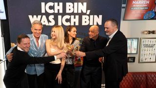 Eine Gruppe von Menschen, sowohl Männer als auch Frauen, steht vor einem Banner und stößt mit Weinläsern an.