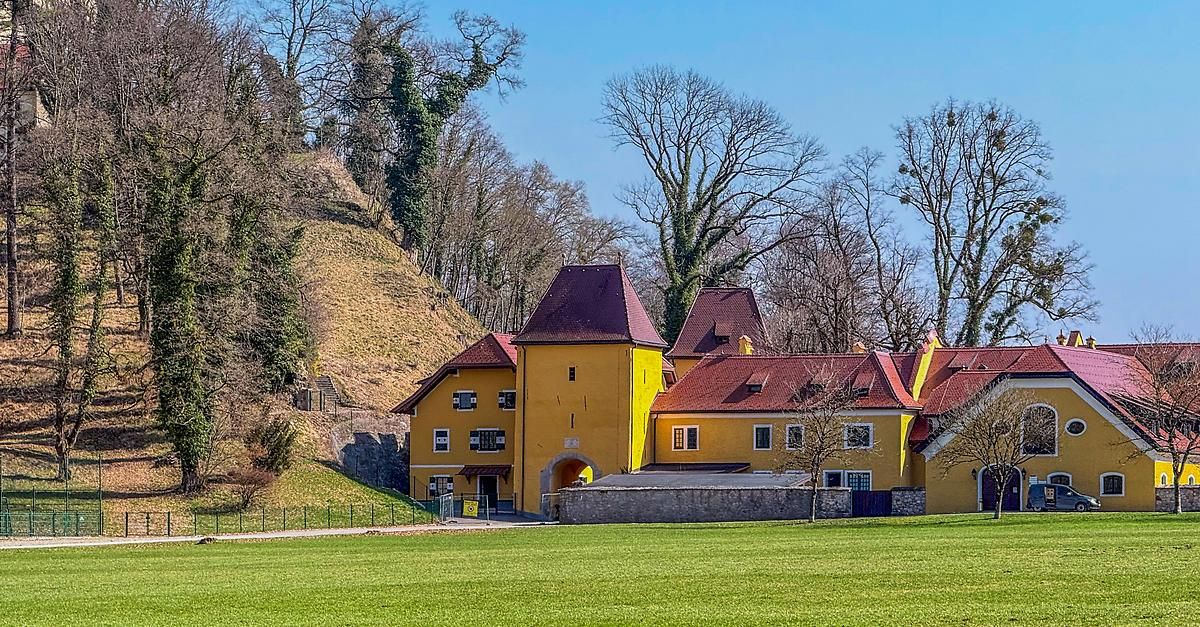 Ein gelbes Schloss mit roten Dächern steht auf einem Hügel, umgeben von grünen Bäumen und Gras unter einem blauen Himmel.