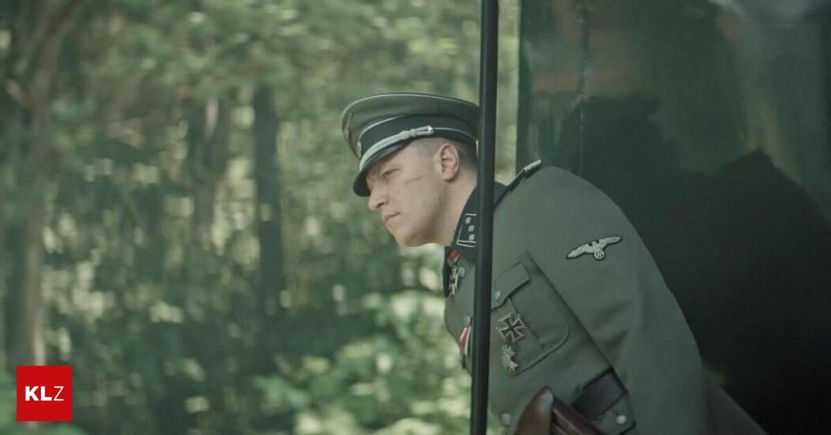 Ein Soldat in einer Militäruniform steht in einem Wald, in Richtung Boden blickend.