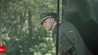 Ein Soldat in einer Militäruniform steht in einem Wald, in Richtung Boden blickend.