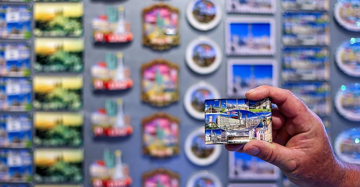 Eine Person hält eine kleine Andenkenkarte mit Bildern von Gebäuden und einer Stadt vor einer Wand mit vielen kleinen Bilderrahmen.