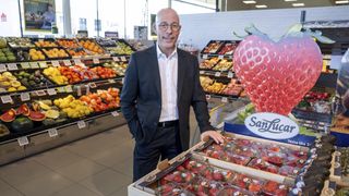 Ein Mann steht lächelnd in einem Lebensmittelgeschäft in der Nähe einer Erdbeer-Ausstellung. Verschiedenes Obst und Gemüse sind im Hintergrund zu sehen.