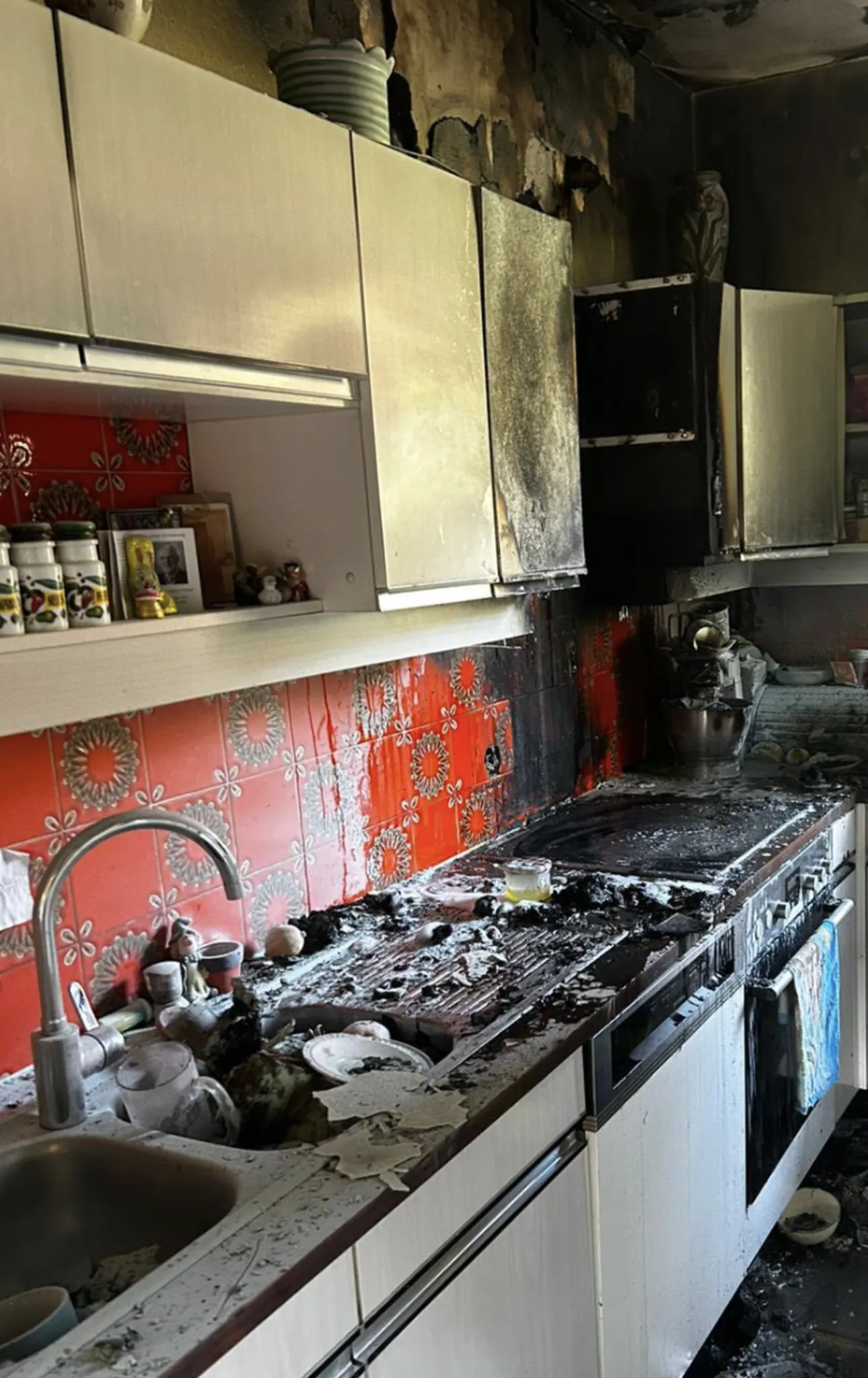 Eine Küche ist zu sehen, mit einem verbrannten Herd und Küchenbereich. Es gibt rote Kacheln und Rußschäden an den Schränken.