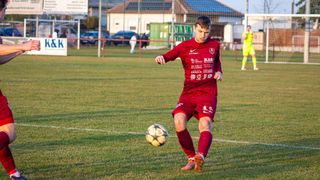 Ein Fußballspieler in roter Uniform tritt einen Ball auf einem Feld, mit einem verschwommenen Hintergrund aus geparkten Autos und einem Gebäude.