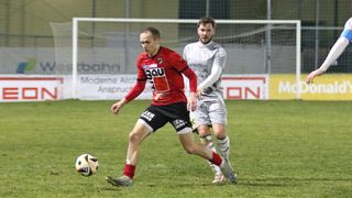 Zwei Fußballspieler sind auf einem Feld. Einer ist in Rot und der andere in Grau. Sie jagen einem Fußball hinterher. Dahinter befindet sich ein Zaun und ein Schild.