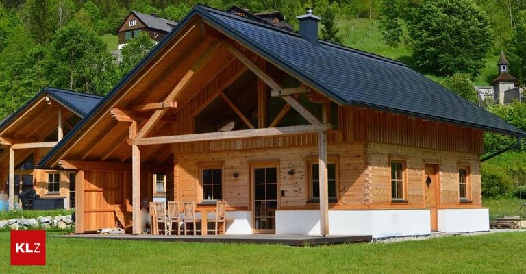 Ein Holzhaus mit schwarzem Dach und Glasfenstern. Eine hölzerne Veranda und ein Tisch mit Stühlen stehen vor dem Haus. Bäume und ein Haus befinden sich hinter dem Haus.
