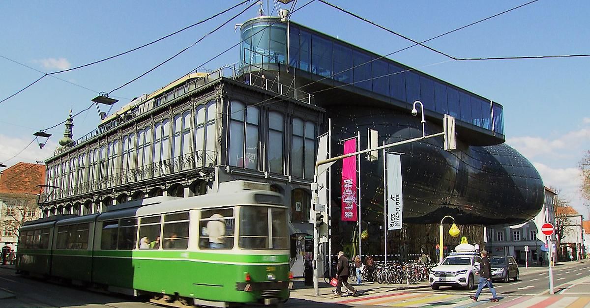 Eine grüne Straßenbahn fährt auf einer Straße vor einem modernen Gebäude mit Glaswänden und einem gebogenen Dach. Menschen gehen auf dem Bürgersteig, und Fahrräder sind in der Nähe geparkt.