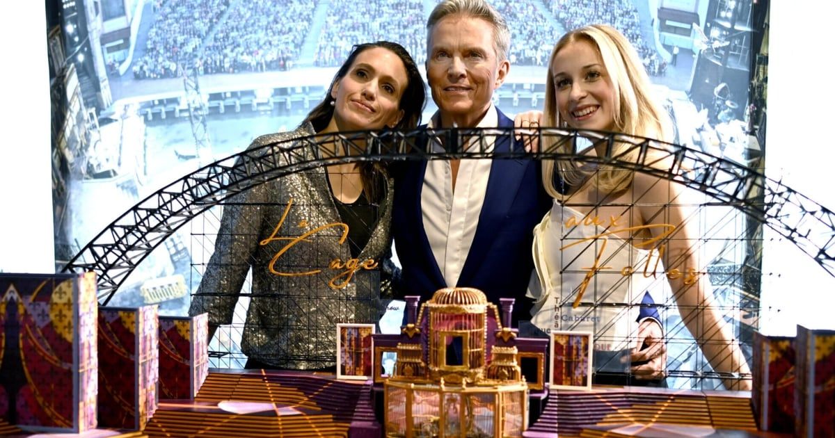 Ein Mann und zwei Frauen stehen vor einem Modell einer Bühne mit einem großen Bogen, einem Vogelkäfig und anderen Dekorationen. Das Modell ist mit 'Euge' beschriftet. Die Frau auf der linken Seite trägt ein Pailletten-Top. Im Hintergrund ist ein Publikum zu sehen.