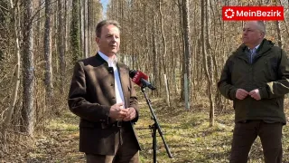 Ein Mann steht in einem Wald und spricht in ein Mikrofon mit dem ORF-Logo. Er trägt einen braunen Mantel und ein weißes Hemd.