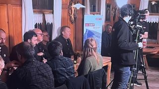 Eine Gruppe von Menschen sitzt um einen Tisch mit einem Schild, das 'ein ORT WORT' besagt. Ein Mann mit einer Kamera steht in der Nähe.