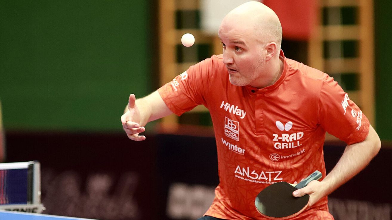 Ein Mann in einem orangefarbenen Shirt spielt Tischtennis und schlägt einen Ball mit einem Schläger. Er sieht den Ball mit einem ernsten Gesichtsausdruck an.